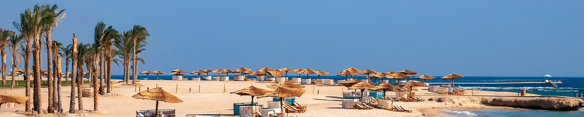 Uitzicht over strand van Port Ghalib met rieten parasollen, ligbedjes en palmbomen in Marsa Alam, Egypte