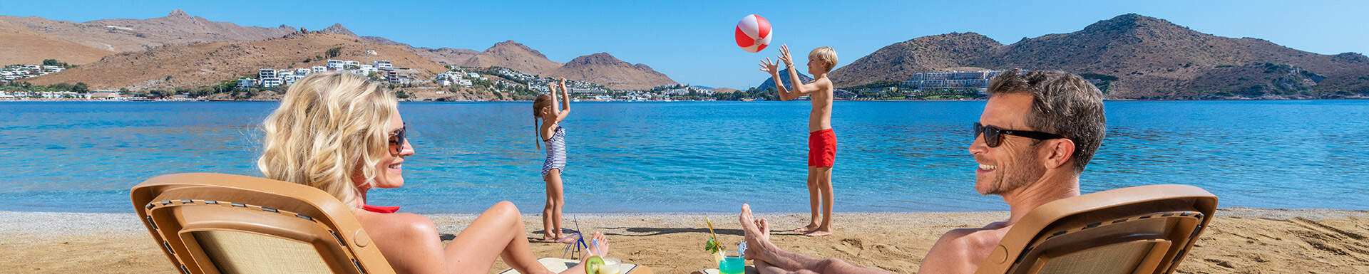 man en vrouw op het strand die lachend naar elkaar kijken met twee kinderen die een bal overgooien naast de zee