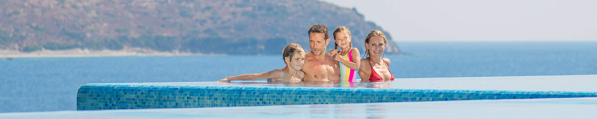 man en vrouw met twee kinderen in badkleding in infinity pool met uitzicht op zee en rotsen