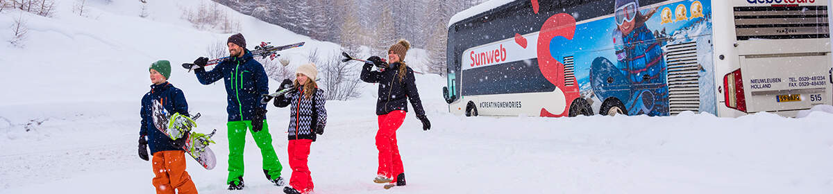 familie loopt met skies en snowboard op schouders vanuit sunweb bus de sneeuw in in skigebied